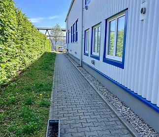 Průmyslový areál s dlážděným chodníkem vedle budovy se světlou fasádou a modrými rámy oken, obklopený zeleným živým plotem.