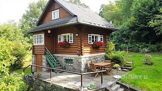 Dřevěná chata s kamennými základy, obklopená zelení, nabízí terasu s venkovním posezením. Tradiční rustikální styl doplňují květinové truhlíky.