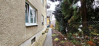 Úzká zahradní cesta se zámkovou dlažbou vedle bytového domu, obklopena keři a stromy, ideální pro klidné procházky a rekreaci