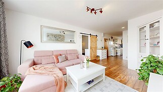 Světlý obývací pokoj s dřevěnou podlahou, moderní pohovkou a velkými okny, které zajišťují dostatek přirozeného světla.