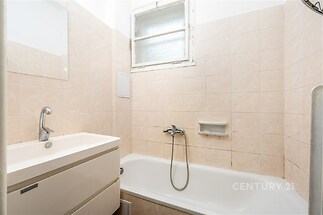 Kompaktní koupelna s vanou, bílými keramickými dlaždicemi a umyvadlem, přirozené světlo skrze matné okno, minimalistický design.