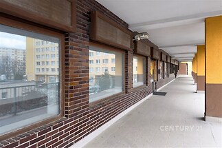 Dlouhá krytá terasa s dlažbou a cihlovým obkladem, velká okna nabízejí výhled na okolní bytové domy, vhodná pro relaxaci a odpočinek.