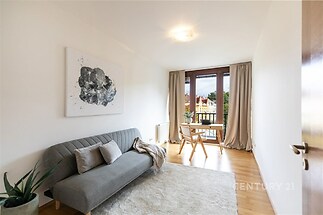 Světlý obývací pokoj s parketovou podlahou, velká okna s výhledem na balkon, moderní nábytek a minimalistická dekorace.