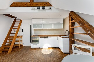 Podkrovní kuchyň s moderním vybavením, dřevěná podlaha a odkryté trámy, světlé skříně a dřevěné schody vytvářejí útulnou atmosféru.