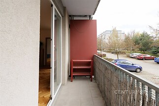 Balkon s dlažbou, výhledem na zeleň a parkoviště, sousedící s parketovým pokojem, nabízí dostatek přirozeného světla pro útulné posezení.