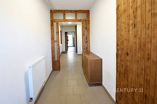 Úzká chodba s dlažbou, dřevěnými dveřmi, bílými stěnami a radiátory. V zadní části vestavěná skříň. Světlý interiér v rustikálním stylu.