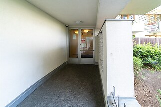 Vstupní chodba s moderním osvětlením a dlážděnou podlahou, vedoucí k proskleným dveřím s bezpečnostním zámkem, zajišťuje bezpečí a komfort.