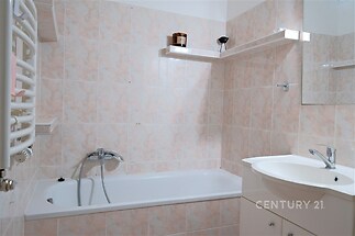 Koupelna s vanou, světlými dlaždičkami a moderním umyvadlem, doplněná úložnými prostory a topným žebříkem.