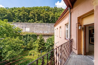 Balkon s dlažbou nabízí výhled na zelené okolí a železniční trať, světlo proniká z velkých oken, a budova má historický styl.