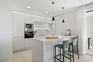 Moderní kuchyň s bílými skříňkami, dvěma závěsnými světly a vestavěnými spotřebiči. Prostorný design s elegantním barem a dlažbou.