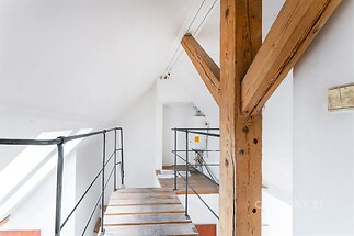 Podkrovní prostor s dřevěnými trámy, moderní kovové zábradlí, světlá úprava stěn, industriální styl, přirozené osvětlení, výhled z velkých oken