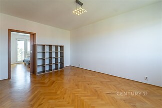Světlý obývací pokoj s parketovou podlahou a moderním osvětlením, vedoucí do ložnice s balkónem, ideální pro rekonstrukci.