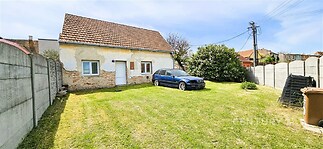 Menší zahrada s travnatou plochou, ohraničená betonovým plotem, s původní přízemní budovou s červenou střechou, vyžadující rekonstrukci.