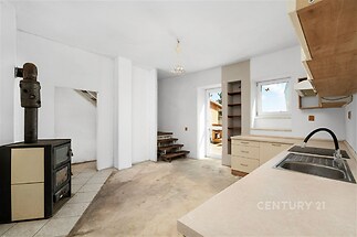 Prostorná kuchyň s dřevěnými policemi, dlažbou a vintage kamny. Velká okna poskytují dostatek přirozeného světla, kuchyň je ideální pro rekonstrukci.