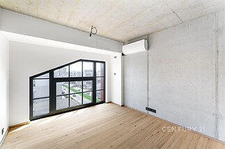 Světlý obývací pokoj s parketovou podlahou a velkými okny, moderní klimatizace a betonové stěny vytvářejí stylový interiér.