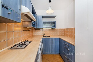 Kompaktní kuchyň s modrými dřevěnými skříňkami, dlaždičkovým obkladem a vestavěnou varnou deskou. Moderní osvětlení zajišťuje světlost prostoru.