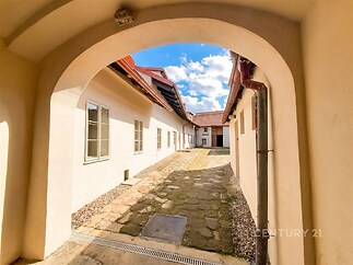 Kamenný dvůr s obloukovým průchodem, dlážděnou cestou a bílými fasádami. Rustikální styl a slunečné počasí dodává prostoru jedinečný charakter.