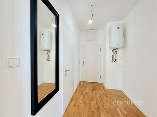 Úzká chodba s parketovou podlahou, bílými stěnami a velkým zrcadlem. Místnost je moderní, nabízí vstupní dveře a nástěnný ohřívač vody.