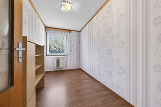 Úzký pokoj s dřevěnou podlahou, dekorativní tapety a velké okno. Pokoj má vestavěné skříně a je vhodný k rekonstrukci.