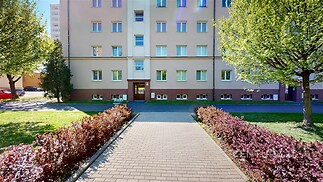 Venkovní fasáda bytového domu se světlou omítkou, upraveným přístupovým chodníkem a okolní zelení, vhodné pro městské bydlení.