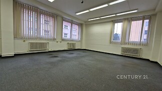 Prostorná kancelář s kobercovou podlahou, velkými okny a vertikálními žaluziemi, vhodná pro moderní pracovní prostředí.