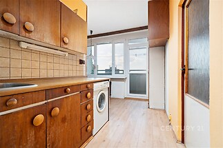 Kuchyň s dřevěnými skříněmi, prostor pro pračku a velkým oknem. Dřevěné prvky a světlá laminátová podlaha dodávají místnosti útulnost.