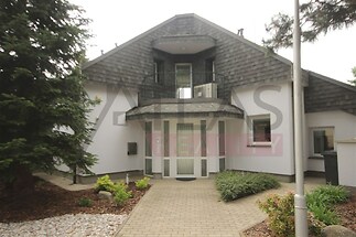 Vchod do rodinného domu s moderní fasádou, střešní tašky a dlážděná cesta, obklopený udržovanou zahradou a jehličnanem.