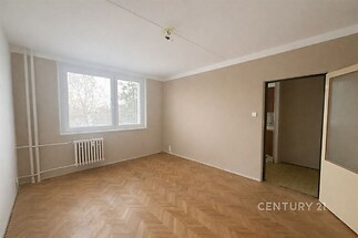 Světlý obývací pokoj s parketovou podlahou, velkými okny a neutrálními stěnami, ideální pro moderní bydlení.