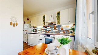 Moderní kuchyň s parketovou podlahou, bílými skříňkami a barevnou dlažbou na zdi. Velká okna poskytují dostatek světla.