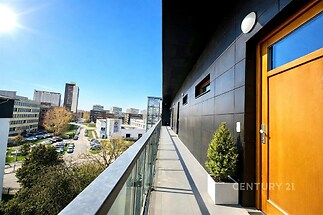 Průchozí balkon s moderním zábradlím a výhledem na město, vedlejší dřevěné dveře a černé obložení budovy.