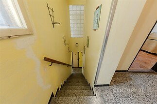 Schodiště s dlažbou, dřevěným zábradlím a velkým oknem, které umožňuje přirozené osvětlení, působí světlým a vzdušným dojmem.