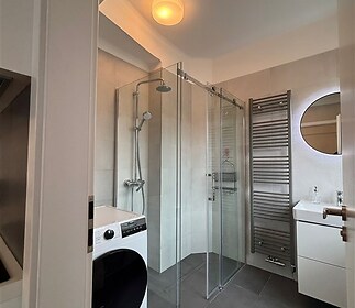 Moderní koupelna s dlažbou, proskleným sprchovým koutem a vestavěnou pračkou, nabízí elegantní design a praktické vytápění s žebříkovým radiátorem.