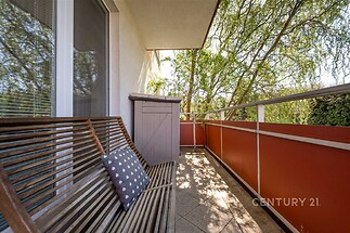 Balkon s dlažbou, dřevěná lavička se vzorovaným polštářem, výhled na zeleň, moderní zábradlí, slunný a klidný prostor vhodný k relaxaci