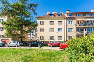 Exteriér bytového domu s udržovanou zahradou, parkováním a červeným autem; ideální lokalita s klidným okolím, vhodná pro rodiny.