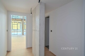 Chodba s dřevěnou podlahou, betonovým pilířem a moderními bílými stěnami, vedoucí do světlého obývacího pokoje s velkými okny.