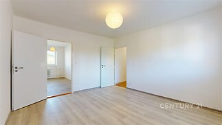 Světlý pokoj s laminátovou podlahou, bílými stěnami a centrálním osvětlením, v bytě s moderním designem a velkou průchodností mezi místnostmi.