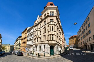 Historická budova na rohu s klasickou fasádou a červenou střechou, nachází se na dlážděné ulici v centru města, dobře udržovaný stav