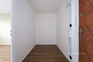 Vstupní chodba s dřevěnou podlahou, čisté bílé stěny a bezpečnostní dveře. Moderní a světlé provedení nabízí praktické využití prostoru.