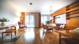 Světlý obývací pokoj s parketovou podlahou, dřevěným nábytkem a velkými okny, které poskytují dostatek přirozeného světla.