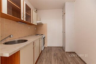 Úzká kuchyň s parketovou podlahou, dřevěnými skříňkami a světlou pracovní deskou, moderní keramická dlažba na zdi kolem sporáku.