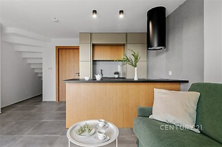 Moderní kuchyň s dřevěnými dvířky, betonová dlažba a světlý obývací prostor. Disponuje minimalistickým designem a otevřeným konceptem.
