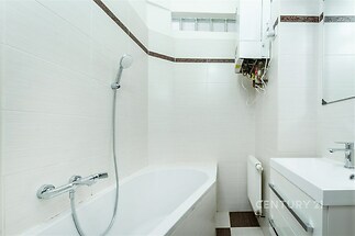 Moderní koupelna s bílými dlaždicemi, vanou a sprchou, závěsná skříňka s umyvadlem, světlé stěny dodávají prostoru vzdušnost.