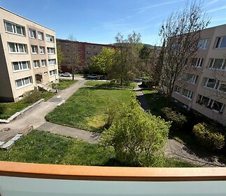 Výhled z balkonu na upravený společný vnitroblok se zelení mezi bytovými domy, světlý sluneční den, okolní bytové domy s klasickými fasádami