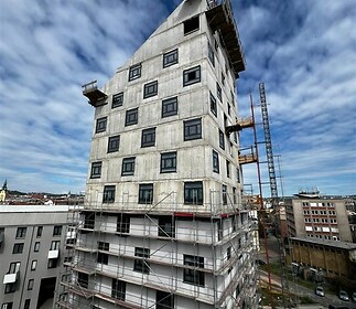 Nově budovaná bytová budova s moderním designem, betonovým povrchem a velkými okny, nacházející se v rozvíjející se městské lokalitě.