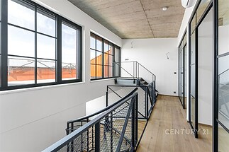 Světlá chodba s velkými okny, moderním průmyslovým stylem a dřevěnou podlahou. Skleněné stěny s kovovými rámy dodávají industriální vzhled.