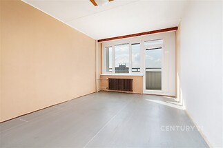 Prázdný pokoj s velkými okny a linoleovým podlahovým povrchem, který umožňuje přirozené světlo; světlý byt v původním stavu.