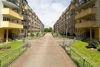 Symetrické bytové domy se světlými fasádami a balkony, vydlážděná promenáda mezi budovami, upravené zelené zahrady a moderní architektura.