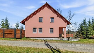 Čelní pohled na moderní rodinný dům s růžovou fasádou, červenou střechou a plastovými okny, obklopený přírodou a oplocením.