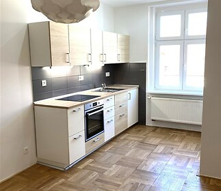 Moderní kuchyň s parketovou podlahou, světlá díky velkým oknům, nabízí dřevěné skříňky a vestavěné spotřebiče v rekonstruovaném stylu.
