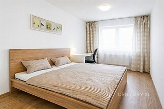 Moderní ložnice s dřevěnou podlahou, velkým oknem a světlým interiérem, vybavená pracovní stůlcem a elegantní dekorací na stěně.